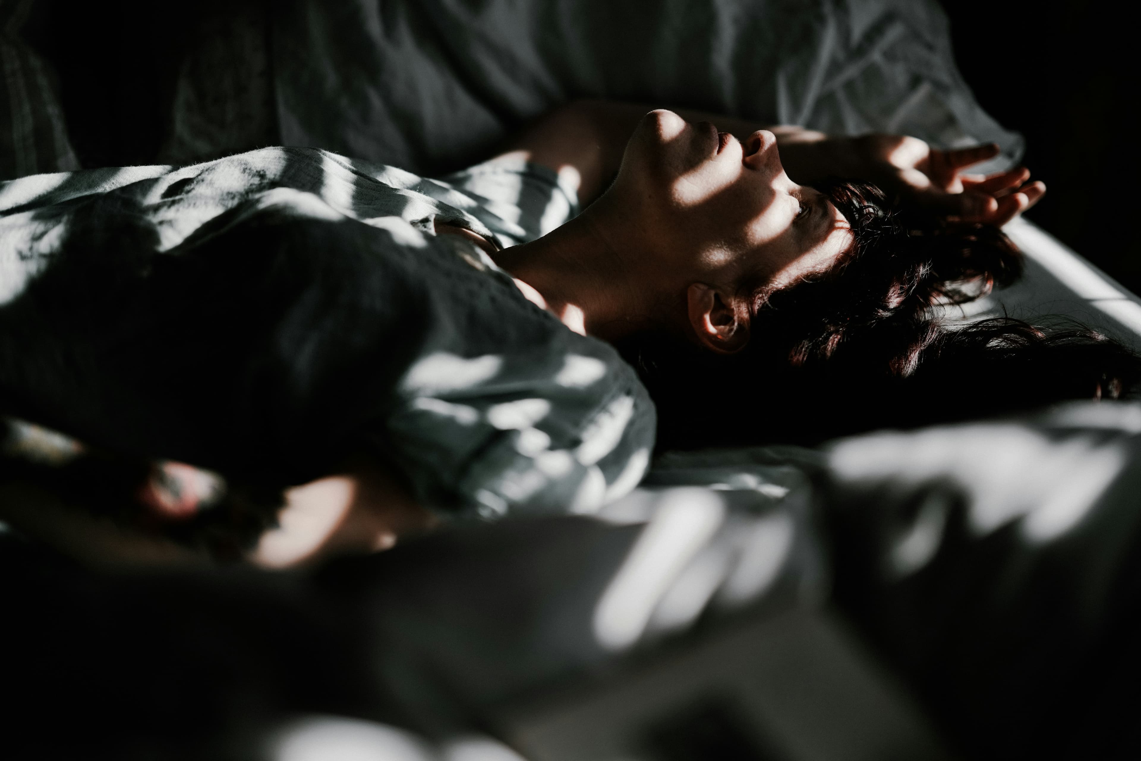 A woman asleep on her back under a duvet, bands of low sun falling across the bed
